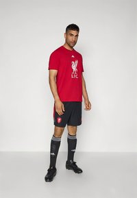 Camiseta roja de manga corta con el logo del Liverpool FC; combinada con pantalones cortos negros con detalles en rojo; calcetas negras con rayas blancas y rojas; botas negras.