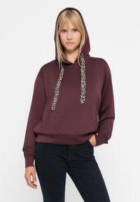 Bordowy dres z przednią kieszenią, z sznurkami w leopardzi wzór i ściągaczami na rękawach. Materiał wydaje się miękki i casualowy.