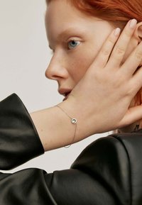 Pulsera de cadena de plata con un acento de gema clara circular, llevada en una muñeca de piel clara, con una manga de cuero negro al fondo.