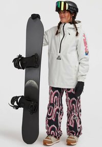 Woman in white jacket, patterned snow pants, and helmet holding a black snowboard with a moon graphic against a plain background.