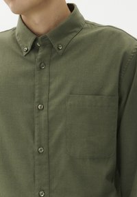 Olivno zelena srajca s gumbi, izdelana iz tkanine z gladko teksturo, z zgornjim žepom, ovratnikom na gumb in ujemajočimi gumbi.