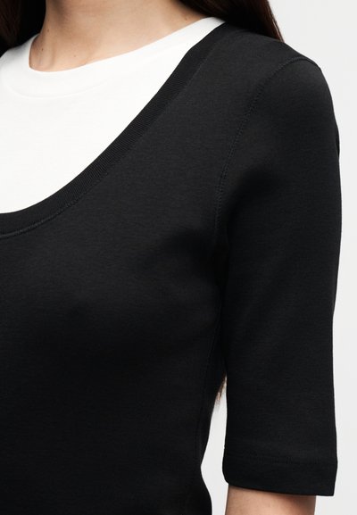 Κοντινό πλάνο ενός ατόμου που φοράει μαύρο τοπ με λαιμόκοψη scoop από πάνω από λευκό μπλουζάκι με στρογγυλή λαιμόκοψη, δείχνοντας τον άνω κορμό και τον ώμο.