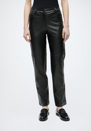 Personne portant un pantalon en simili cuir noir taille haute et des mocassins noirs brillants, debout devant un fond uni.
