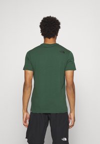 Grön kortärmad t-shirt i bomull, med rund halsringning och en liten svart logotyp på övre ryggen, tillsammans med svarta shorts.