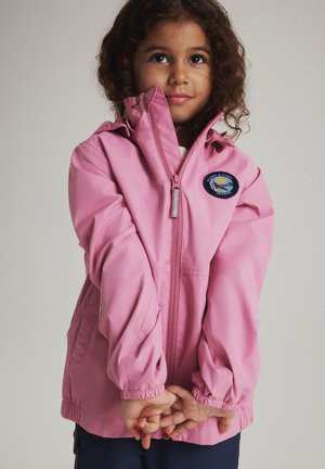 Jong meisje met krullend haar, gekleed in een roze jas met rits en capuchon, staand met de handen gevouwen voor zich.