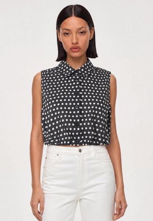 Kvinde med glat mørkt hår iført en ærmeløs sort bluse med hvide prikker og hvide bukser med højt liv, stående mod en neutral baggrund.