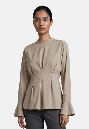 Donna che indossa una camicetta beige a maniche lunghe con polsini svasati e pieghe in vita, abbinata a pantaloni grigio scuro su sfondo bianco.