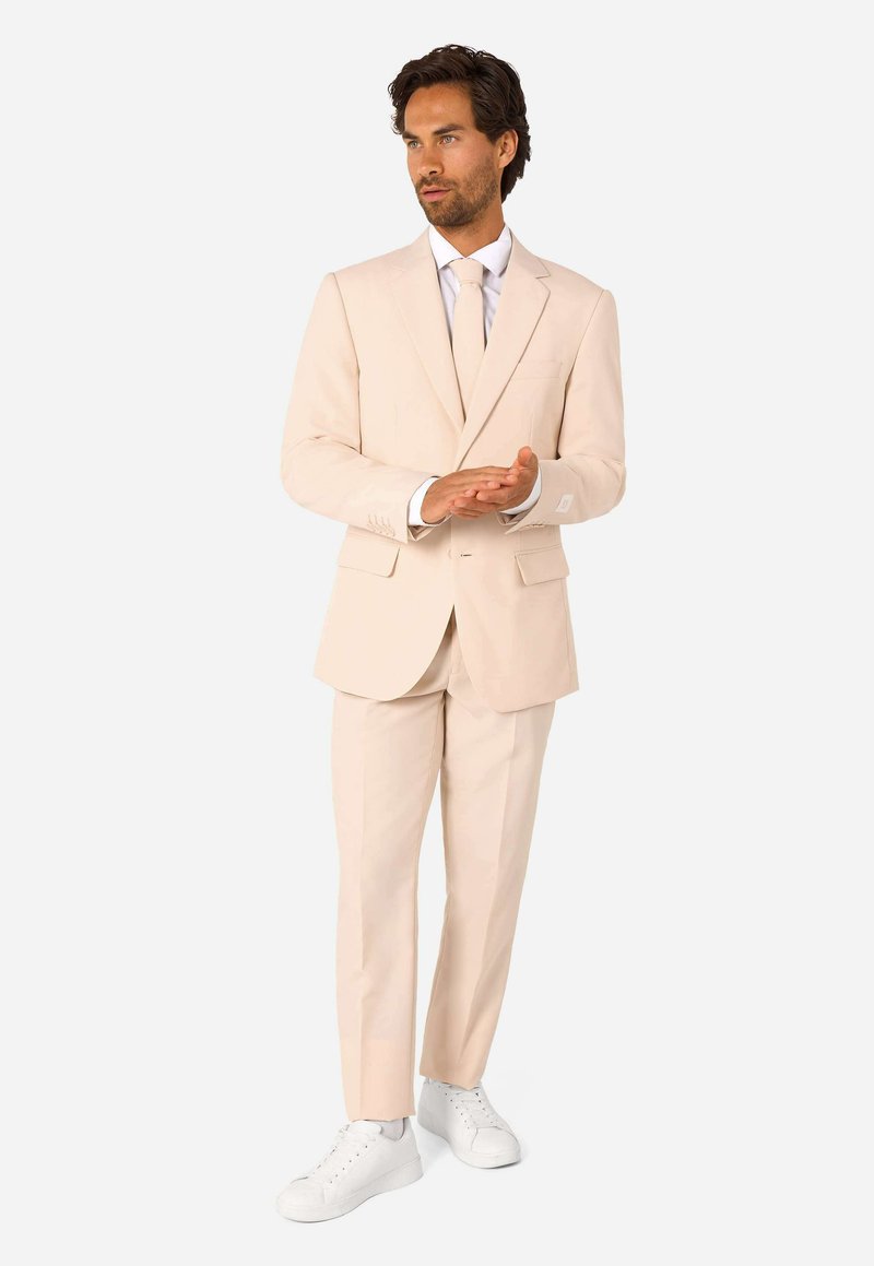 Man standing wearing a beige suit, white shirt, beige tie, and white sneakers, with hands clasped and looking to the side.