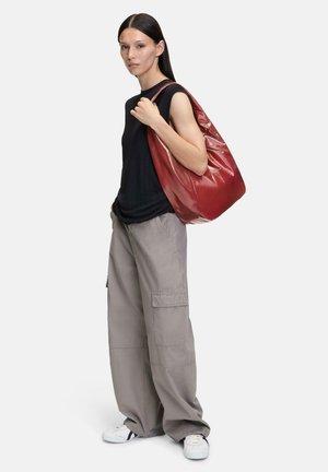 Rote, glänzende Schultertasche aus synthetischem Material, mit abgerundeter Form und weicher Textur, mit einer Hand gehalten.