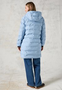 Ljusblå puffjacka med huva, med quiltad textur och knälång design. Matchad med blå jeans och bruna skor.