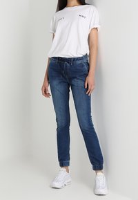 Camiseta blanca de algodón con mangas remangadas, con texto negro "NO ESPERES." Combinada con pantalones jogger de mezclilla azul y zapatillas blancas.