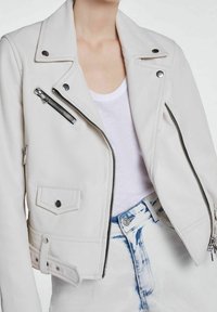 Biała kurtka motocyklowa z imitacji skóry z wyciętym kołnierzem, kieszeniami na suwak, ozdobnymi guzikami i krótkim krojem, w zestawieniu z białym topem.