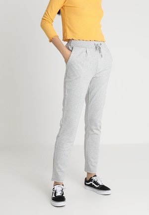 Frau trägt hellgraue Jogginghosen mit Kordelzug, schwarz-weiße Sneakers und ein gelbes, langärmeliges, bauchfreies Oberteil, mit der Hand in der Tasche.