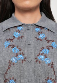 Grijs gebreid cardigan met een kraag. Bevat blauwe en bruine floral borduursels, drie knopen aan de voorkant, ribbelstructuur.