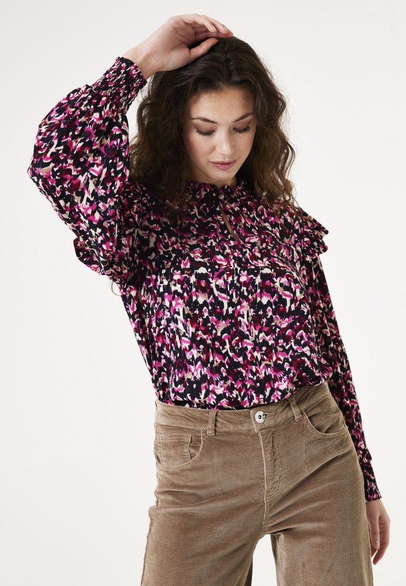 Langdoorshirt in een zwarte stof met een roze en witte abstracte print, met een gestructureerde ruffle halslijn en knoopdetails. Gecombineerd met beige corduroypantalons.