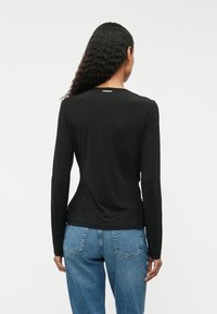 Melnas garām piedurknēm krekls, izgatavots no gludas auduma, ar noapaļotu kakla izgriezumu un smalku logotipa detaļu aizmugurē. Apvienots ar ziliem džinsiem.