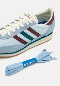 Ljusblå mockasko med vinröda och teal-ränder, cremefärgade snören och en beige sula. Inkluderar en blå skosnöre med varumärkeslogotyp.