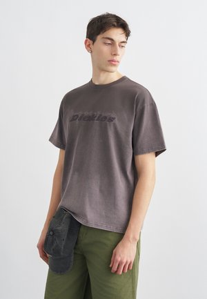 Νεαρός άνδρας με φθαρμένο σκούρο γκρι μπλουζάκι Dickies και χακί παντελόνι, κρατώντας μαύρο καπέλο στο αριστερό του χέρι.