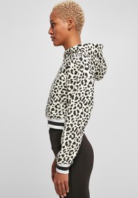 Skračená mikina z béžového materiálu s leopardím vzorom. Obsahuje kapucňu so šnúrkou a čierne manžety s bielymi pruhmi.