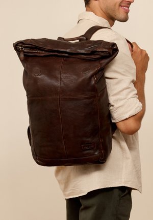 Sac à dos en cuir marron avec un haut roulé, des bretelles ajustables et des détails de couture visibles. Présente un patch avec le logo de la marque sur le devant.