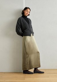 Cardigan grigio, dolcevita nero, gonna maxi in satin oliva e ballerine nere texturizzate, su uno sfondo grigio chiaro. Pavimento in legno visibile.