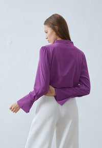 Lilla bluse med høy krage, puffede ermer og flagrende mansjetter. Laget av glatt stoff, brukt med hvite vide bukser.