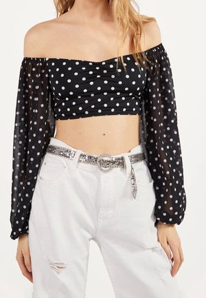 Vrouw draagt een zwarte off-shoulder crop top met witte stippen en doorschijnende mouwen, gecombineerd met witte gescheurde spijkerbroek en een zilveren paillettenriem.