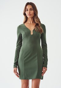 Robe verte ajustée en matière extensible, dotée de manches longues, d'un décolleté en V profond et d'une fente sur le côté. Texture lisse avec des détails de couture.