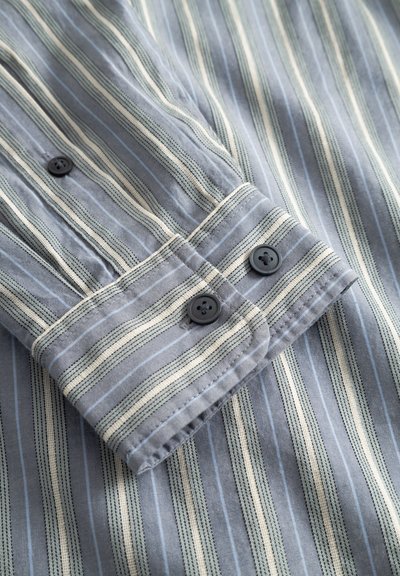 Siva srajca z vzorcem izmenjujočih se tankih in debelejših pokončnih črt, črni gumbi na manšetah in gladka tekstura tkanine.
