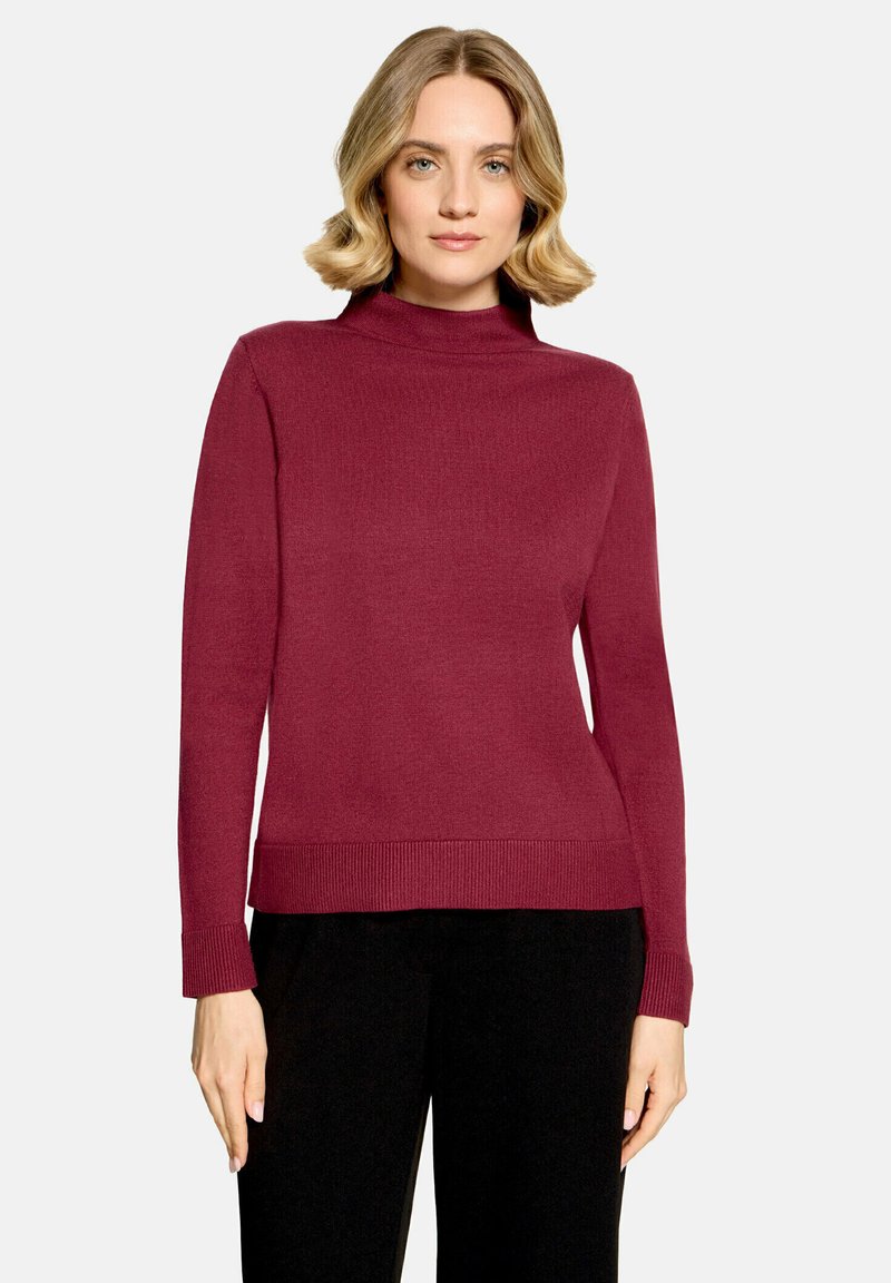 Femme aux cheveux blonds portant un pull bordeaux à manches longues et col montant, et un pantalon noir, debout devant un fond uni.