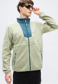 Φωτεινό πράσινο φλις μπουφάν με μπροστινό φερμουάρ σε τυρκουάζ, με πλαϊνές τσέπες και αντίθετες λεπτομέρειες φερμουάρ. Το μοντέλο φορεί γυαλιά ηλίου.