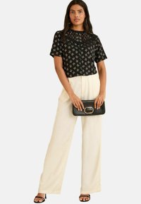 Haut noir à manches courtes avec des motifs de nœuds blancs, associé à un pantalon large beige clair. Comprend une pochette noire avec un accent doré.