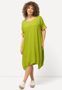 Zelené oversized šaty s krátkými rukávy, oranžovými akcenty na manžetách, volný střih a zaoblený lem. Nošeny s bílými platformovými botami.