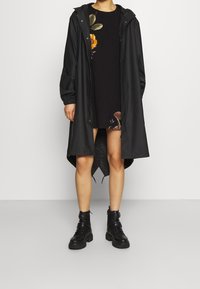 Czarny wodoodporny płaszcz przeciwdeszczowy z kapturem ze sznurkiem, z elastycznymi mankietami i dłuższym tyłem, noszony na czarnej sukience z haftem kwiatowym.