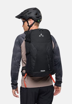 Personne portant un casque de vélo noir et des lunettes de soleil, avec un grand sac à dos noir muni d'un logo réfléchissant et de sangles orange.