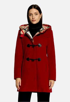 Vrouw draagt een rode duffelcoat met zwarte sluitingen en een geruite capuchonvoering over een zwarte coltrui.