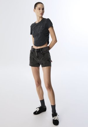 Camiseta negra cropped con texto en relieve, combinada con pantalones cortos de mezclilla negros de cintura alta con un patrón de rayo. Zapatillas negras y calcetines grises.