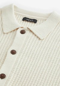 Cremfarbener strukturierter Strickpolo mit gefaltetem Kragen und drei braunen Knöpfen auf der Vorderseite. Das Markenlabel lautet "NEXT."