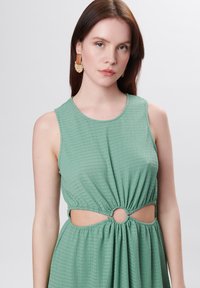 Vestido verde sin mangas con tejido texturizado, que presenta un corte circular en la cintura y un detalle fruncido, combinado con pendientes de colores claros.