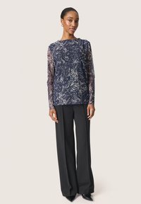 Bluză cu mâneci lungi de culoare bleumarin, cu un model abstract de spirale albe; mâneci transparente și corp solid; asortată cu pantaloni negri largi și pantofi negri cu vârful ascuțit.