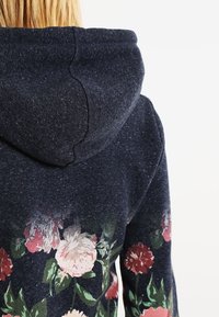Sudadera con capucha en tejido azul marino oscuro, con estampado floral en rosa, rojo y verde en la parte baja de la espalda, con una superficie texturizada.