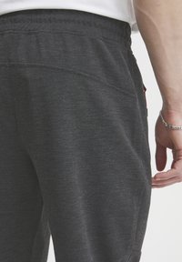 Primer plano de una persona que lleva pantalones texturizados de color gris oscuro y una pulsera plateada en la muñeca derecha, contra un fondo blanco.