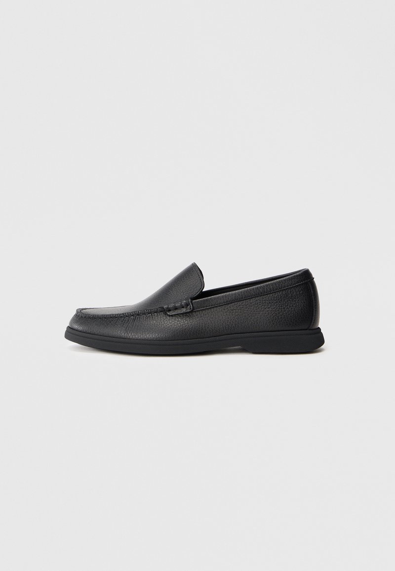 Mocasín negro de cuero, sin cordones, con parte superior texturizada, punta redondeada, tacón bajo y suela de goma, mostrado en perfil lateral sobre un fondo blanco.