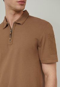 Braunes Polo-Shirt mit Reißverschlusskragen, kurzen Ärmeln und einer aufgesetzten Tasche am Ärmel; aus strukturiertem Material gefertigt.