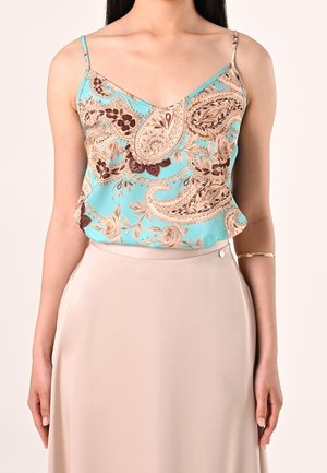 Kvinde iført en turkis ærmeløs top med paisleymønster og beige højtaljet nederdel samt guldbangle på højre arm.