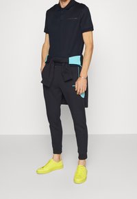 Polo marine, pantalon de jogging noir avec des accents turquoise et baskets jaunes vives. Le tissu semble lisse, coupe décontractée, avec un logo.