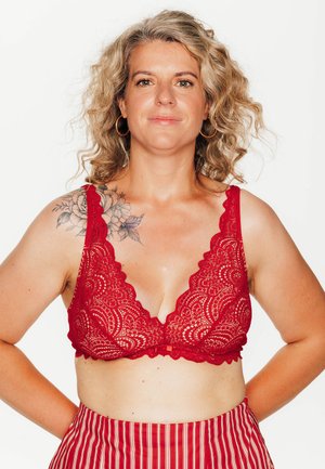 Bralette in pizzo rosso con bordi scallop, scollatura a V profonda e spalline larghe. Tatuaggio floreale significativo sulla spalla sinistra. Gonna a vita alta a righe rosse e bianche.