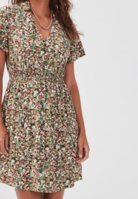 Femme portant une robe florale courte avec des fleurs vertes, blanches, rouges et jaunes, décolleté en V, manches courtes, et un collier en chaîne dorée.