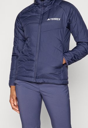 Osoba nosząca granatową puchową kurtkę Terrex z zamkiem błyskawicznym i dopasowane granatowe spodnie, jedna ręka w kieszeni kurtki, stojąca na jednolitym tle.