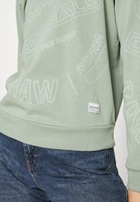 Sudadera verde con patrones de logotipos bordados en blanco, cuello redondo, puños acanalados y una pequeña etiqueta de marca en el dobladillo, combinada con jeans azules.